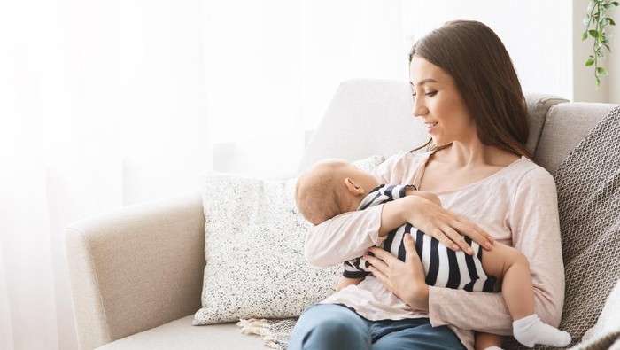 Mother's lullaby. Beautiful mom lulling newborn baby while sitting on sofa at home