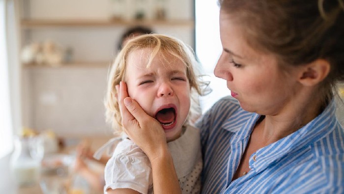 A mother holding a crying toddler daughter indoors in kitchen when cooking.