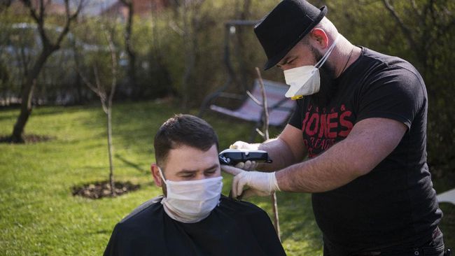 Kosovo hairdresser Driton Kameri (R) wears a facemask as he cuts the hair of a client in his garden on March 30, 2020 in Pristina during the time of COVID-19 pandemic. - In Pristina, a barber is defying coronavirus edits, such as the ban on opening non-essential shops, to practice his art in the open air, but not without first spraying his customers with disinfectant from head to toe. (Photo by Armend NIMANI / AFP)