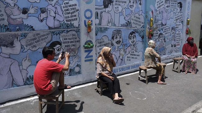 Warga berjemur dengan latar belakang mural (lukisan dinding) komik antihoaks di Kampung Hepi, Joho, Manahan, Solo, Jawa Tengah, Selasa (7/4/2020). Mural tersebut dibuat warga setempat untuk mengedukasi warga tentang hidup bersih dan sehat sebagai upaya mencegah penyebaran COVID-19 sekaligus mengampanyekan gerakan antihoaks. ANTARA FOTO/Maulana Surya/foc.