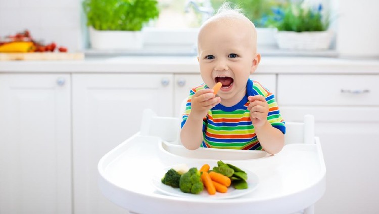 1009591678 Cute baby eating vegetables in white kitchen. Infant weaning. Little boy trying solid food, organic broccoli, cauliflower, carrot and green peas. Healthy nutrition for kids. Child biting carrot.