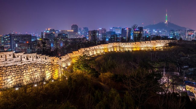 The old city wall lit up in evening as the lights of downtown Seoul shine in the distance.