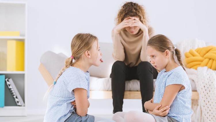 Young pouty sisters arguing with arms crossed and their stressed mom sitting on a sofa in the background