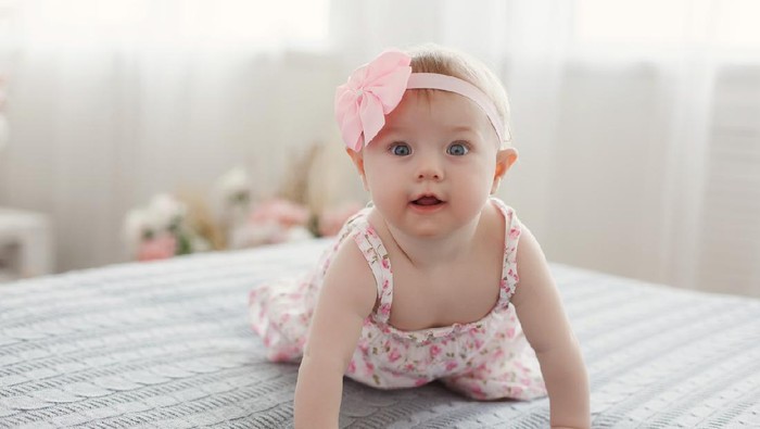Charming beautiful lovely small female baby with plump cheeks creeps on bed, looks innocently on something, being naughty. Cute baby infant plays alone. Newborn kid crawls. Enthusiastic child