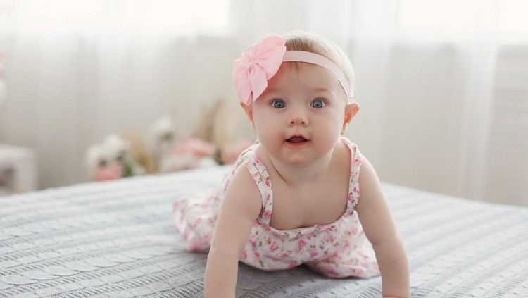 Charming beautiful lovely small female baby with plump cheeks creeps on bed, looks innocently on something, being naughty. Cute baby infant plays alone. Newborn kid crawls. Enthusiastic child