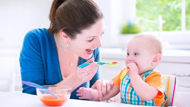 Young attractive mother feeding her cute baby son, giving him his first solid food, healthy vegetable pure from carrot with a plastic spoon sitting in a white sunny kitchen at a window