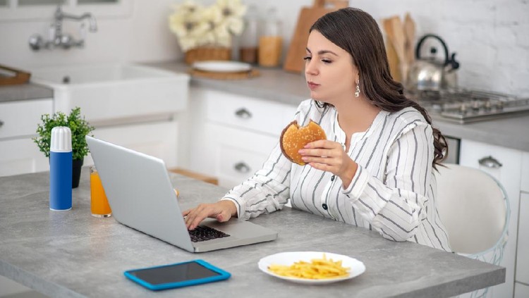 Eating. Dark-haired woman in a striped blouse eating a burger and working