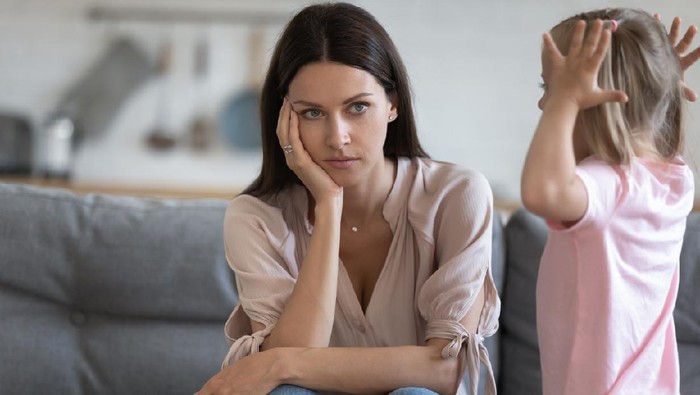 Unhappy mother having problem with noisy naughty little daughter screaming, demanding attention, child tantrum manipulation concept, tired thoughtful mum holding head, sitting on couch at home