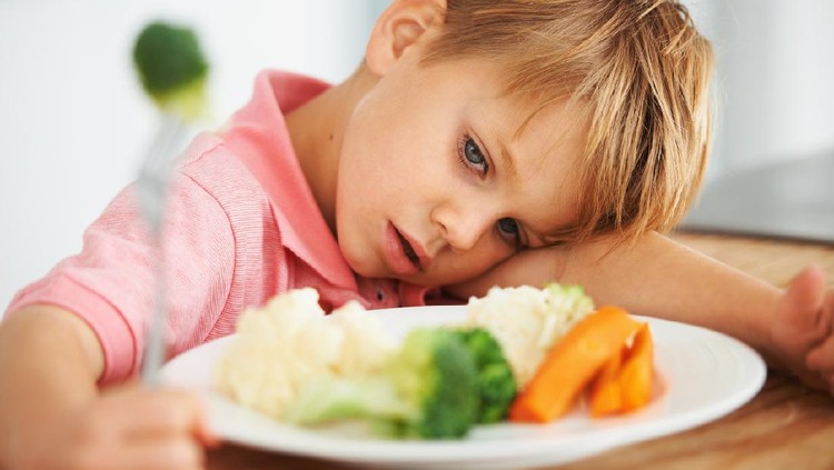 A cute young boy looking at the vegetables on his table while holding a piece of broccoli on his fork