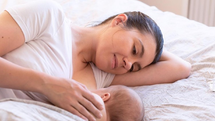 menyusui Beautiful mom lying at the back of the baby while the infant is sleeping on the white bed. Asian mother looking at the baby which is her child with love and care.