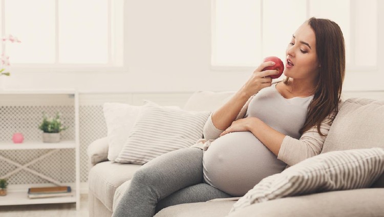 Pregnant woman eating apple at home copy space. Beautiful expectant lady sitting on sofa and having fresh snack. Healthy nutrition and pregnancy concept