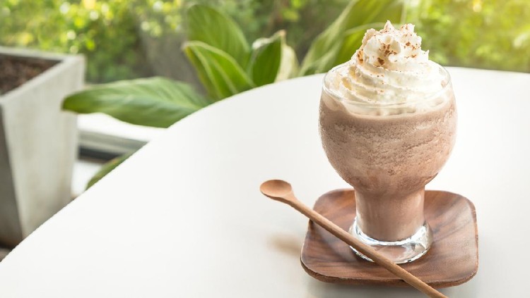 Chocolate smoothies (Cocoa blended) topped with whipped cream and cocoa powder in glass cup on white table. Wooden saucer and spoon. Interior coffee shop.