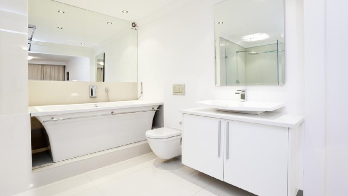 An all white bathroom showing big mirrors, bath, toilet, basin and storage.
