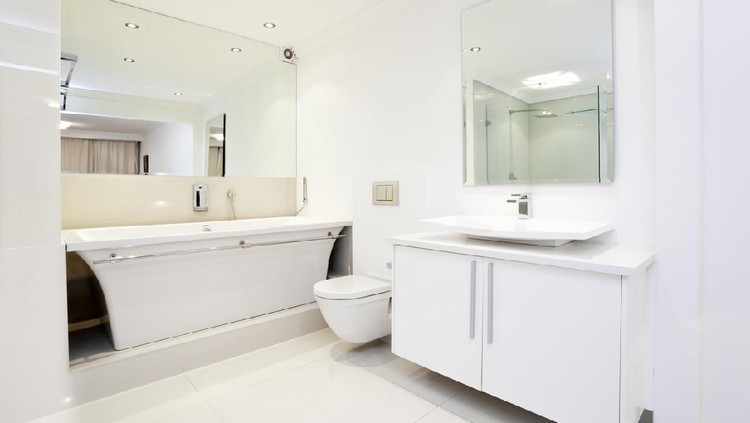 An all white bathroom showing big mirrors, bath, toilet, basin and storage.