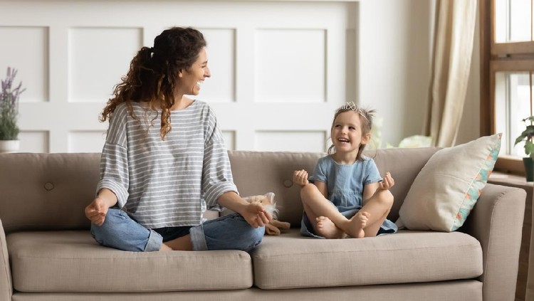 Full length front view smiling young curly mother sitting on comfortable couch with cute playful little preschool daughter in lotus position. Happy mommy practicing yoga exercised with small child.