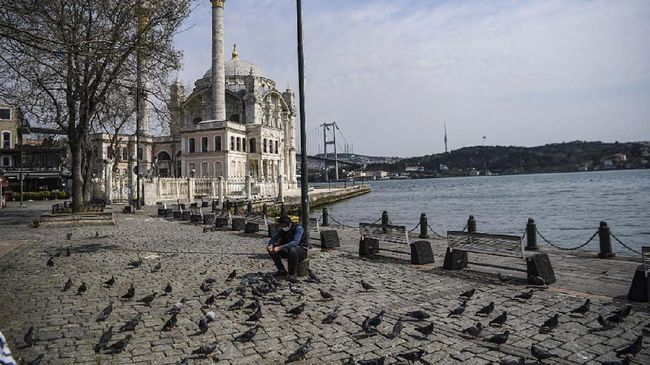 A Turkish police feeds pigeons as July 15 Martyrs' Bridge, known as the Bosphorus Bridge and Ortakoy mosque are seen in the backround in Istanbul on April 11, 2020, as Turkish government announced a two-day curfew to prevent the spread of the epidemic COVID-19 caused by the novel coronavirus. (Photo by Ozan KOSE / AFP)