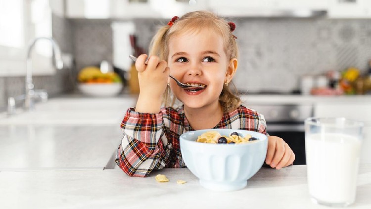 Beautiful little girl having breakfast with cereal, milk and blueberry in kitchen