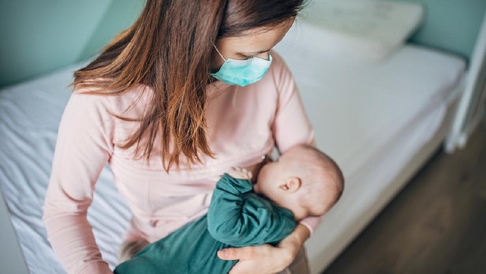 Two people, mother with protective mask breastfeeding her baby son at home, they are at home do to pandemic outbreak quarantine.