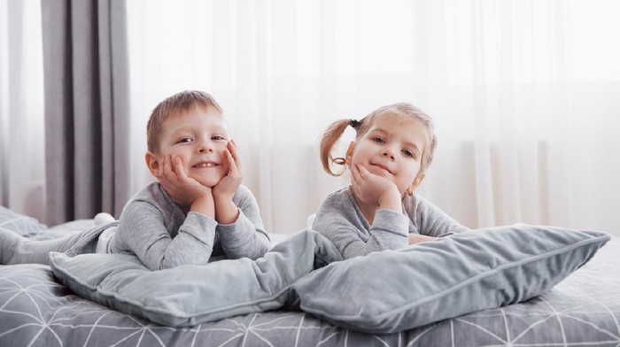 Happy kids playing in white bedroom. Little boy and girl, brother and sister play on the bed wearing pajamas. Nursery interior for children. Nightwear and bedding for baby and toddler. Family at home.
