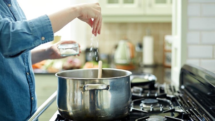 Midsection image of woman adding salt in cooking pot. Utensil is placed on gas stove. Close-up of female seasoning her dish. She is preparing food in domestic kitchen.