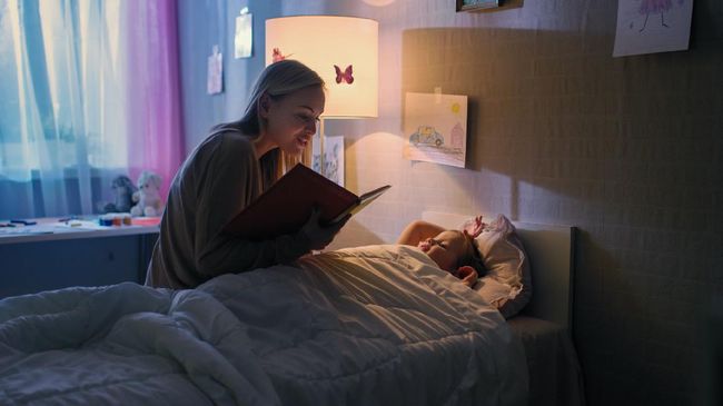 Young Loving Mother Reads Bedtime Stories to Her Little Beautiful Daughter who Goes to Sleep in Her Bed.