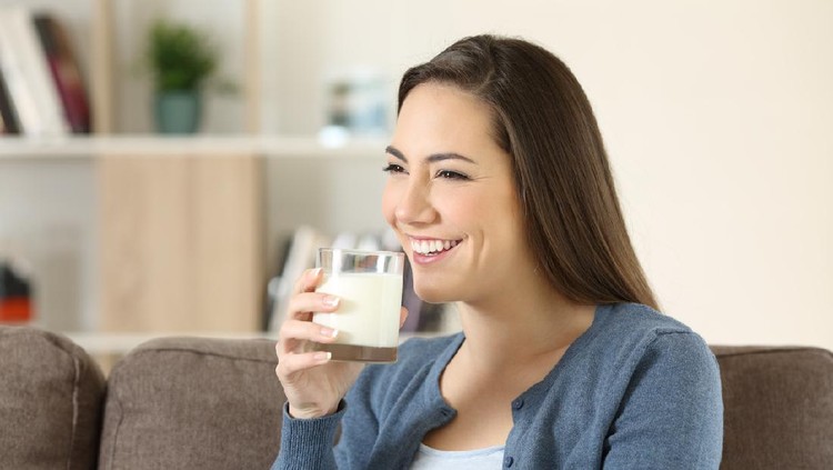 Happy woman holding a glass of milk sitting on a couch in the living room at home
