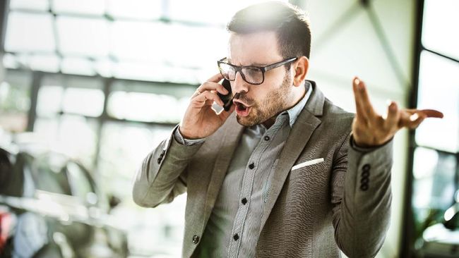 Young displeased businessman arguing with someone over smart phone while being in a car showroom.