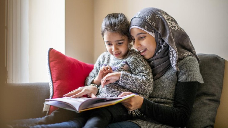1062159252 A pre-teen girl wearing a hijab sits on a couch with her little sister on her lap and reads her a bedtime story. Her sister is engaged in the story.