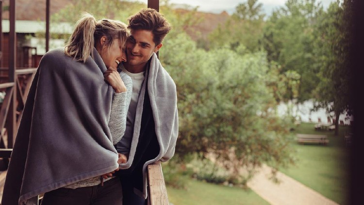 Loving man and woman standing in their hotel room balcony wrapped in blanket. Romantic couple in a blanket standing together on a winter holiday.