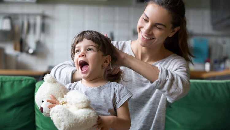 Smiling young loving mum making ponytail to little preschool laughing daughter with toy sitting on sofa, couch in living room at home, mother helping child with hairstyle, nanny caring of pupil