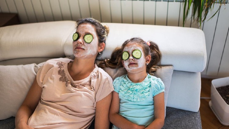 Beautiful mother and daughter with facial mask and slices of cucumber on their eyes enjoying lazy virtual spa day during COVID-19 isolation.