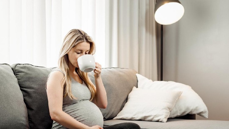Pregnant woman sitting on her sofa drinking coffee and tea.
