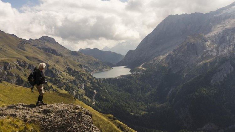 Senior man hiking along hills opposite to the Marmolada in autumn. In distance lake Fedaia. Dolomites, Northern Italy.