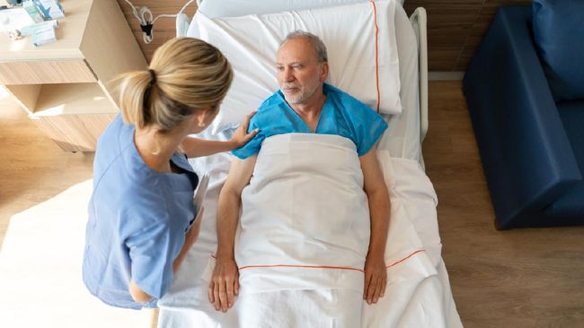 Beautiful nurse comforting senior hospitalized patient lying down on bed talking to him - Healthcare concepts