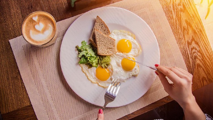 Top view of female hands holding fork and knife while having tasty breakfast at modern european cafe. Young woman eating fried eggs with toasts and morning coffee at restaurant