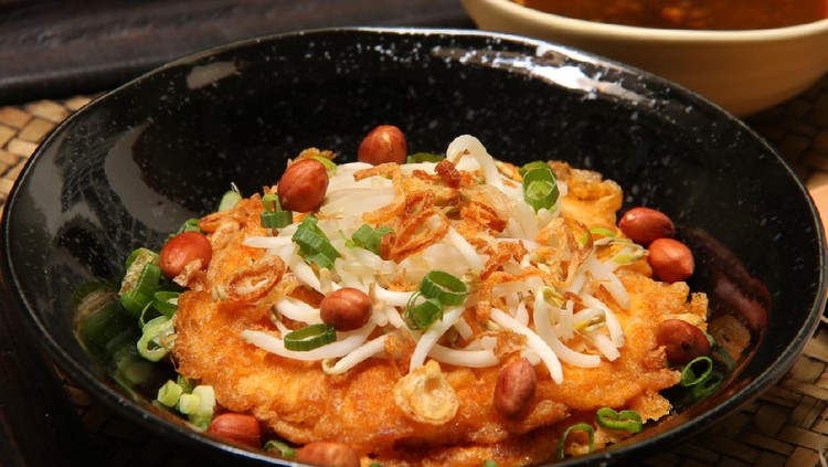 Tahu Telur, a popular egg dish in Surabaya, East Java. Served on black ceramic plate and topped with beansprouts, peanuts, and scallion. Accompanied with spicy soy sauce aside. This is a close-up view of the omelet dish.