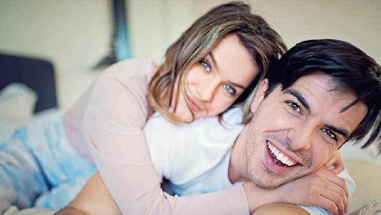 Side view of excited man is lying over woman in bed. They are looking at each other eyes and laughing