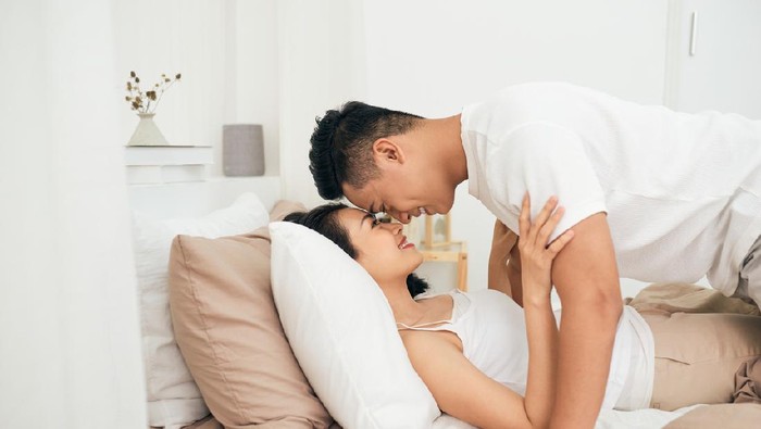 Side view of excited man is lying over woman in bed. They are looking at each other eyes and laughing