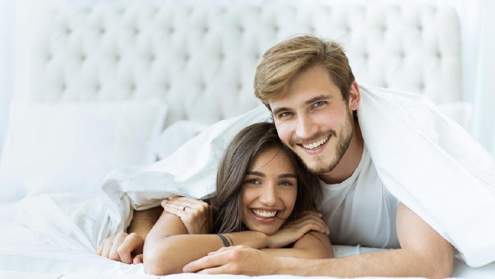 Side view of excited man is lying over woman in bed. They are looking at each other eyes and laughing