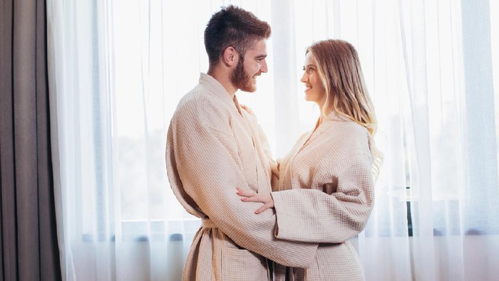 Side view of excited man is lying over woman in bed. They are looking at each other eyes and laughing