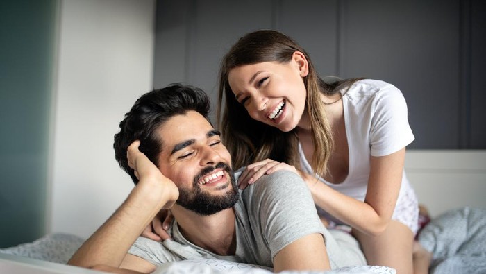 Side view of excited man is lying over woman in bed. They are looking at each other eyes and laughing