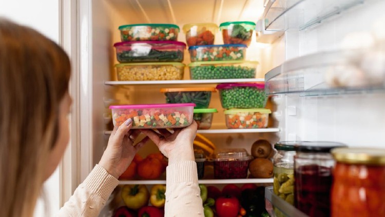 Close up shot of woman taking container with frozen mixed vegetables from refrigerator while looking at camera.
