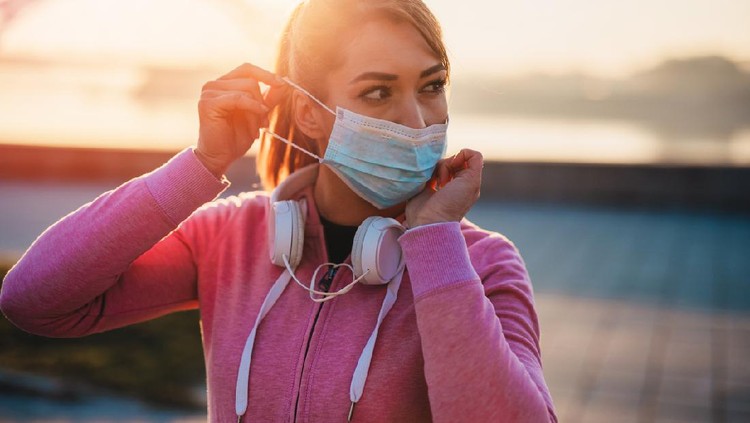 Beautiful female jogger putting protective mask on her face to protect herself from virus or allergy infection. Sunset in background.