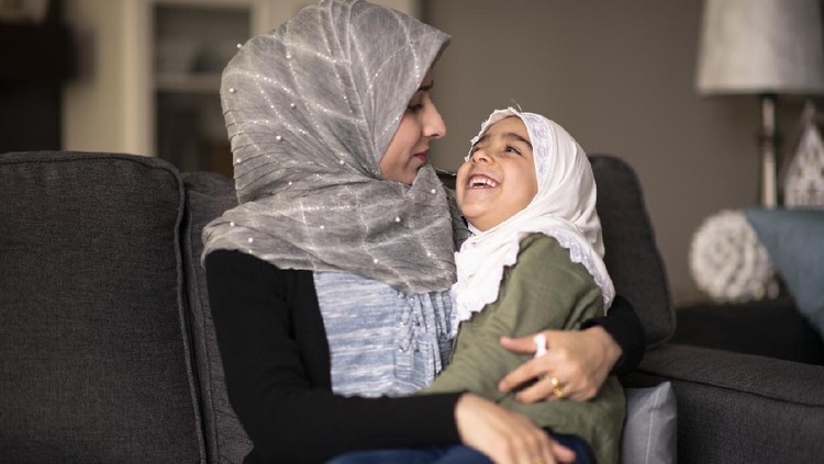 A Muslim mother is smiling while sitting on the couch, with her daughter in her lap. They are both wearing headscarves while cuddling each other.
