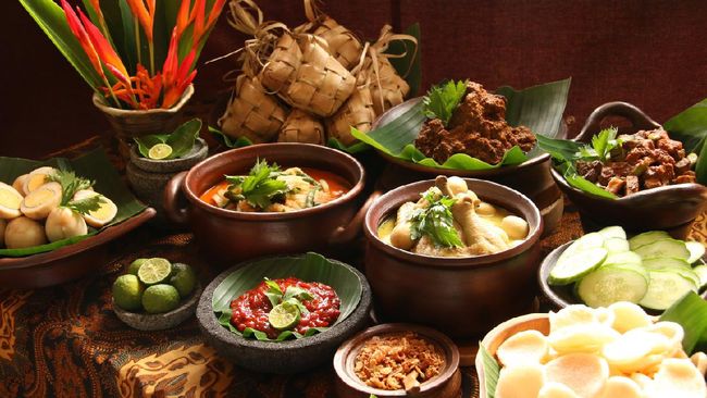 Ketupat Lebaran, the traditional celebratory dish of rice cake with rich side dishes, commonly serving during Eid celebrations. The common side dishes are Beef Rendang (Beef Dry Stew), Beef Liver in Spicy Gravy, Spicy Vegetable Soup, Chicken Curry Soup, and Marbled Soy Egg; while the condiments are fried shallots, shrimp crackers, and red chili sauce. Each dish is plated individually and arranged buffet style on a table; ready to be plated for serving. A vase of decorative banana flowers is used to decorate the table.