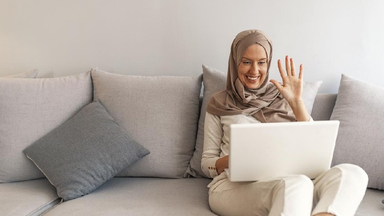 Young Muslim woman having video call via laptop at home. Happy smiling Muslim woman sitting on sofa, couch and using laptop at living room at home, learning language, video calling.