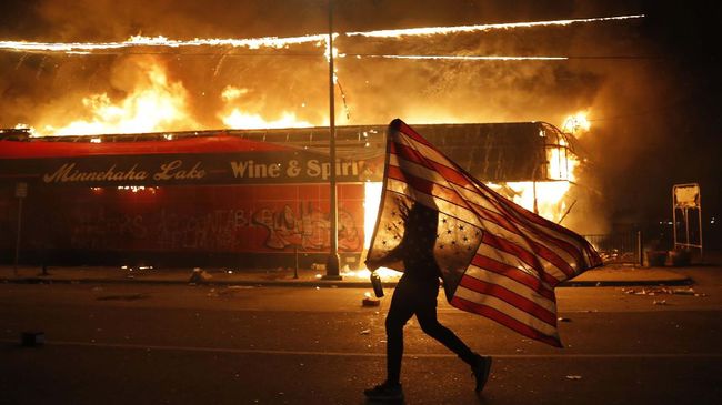 A protester carries the carries a U.S. flag upside, a sign of distress, next to a burning building Thursday, May 28, 2020, in Minneapolis. Protests over the death of George Floyd, a black man who died in police custody Monday, broke out in Minneapolis for a third straight night. (AP Photo/Julio Cortez)