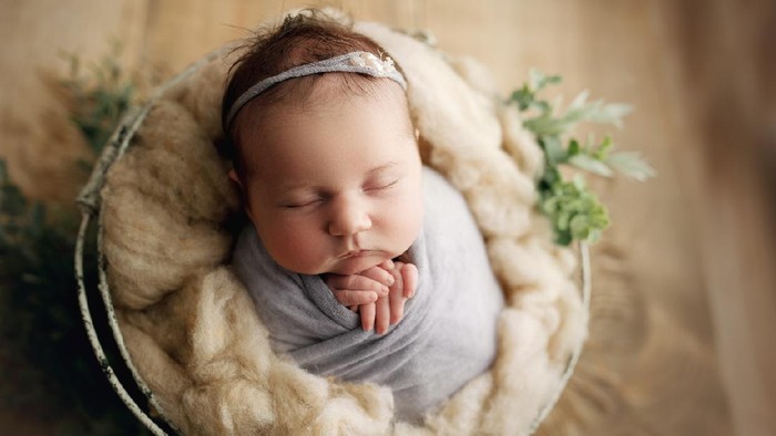 20 day old baby girl with chunky cheeks, swaddled in pale purple with a matching headband, in a prop basket for her newborn milestone photos