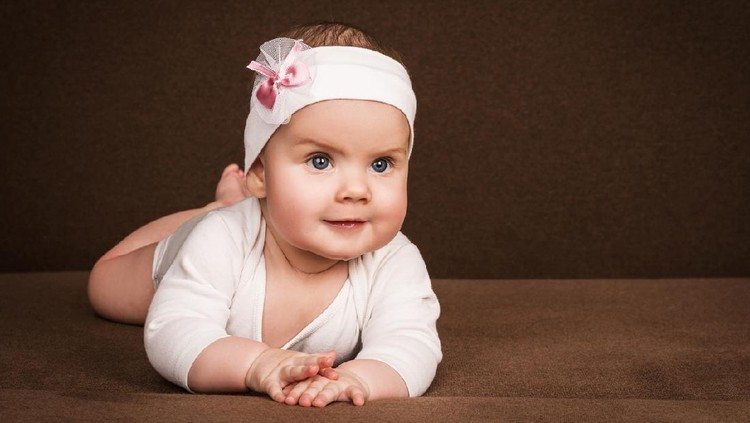 932150536 cute little baby girl with bow flower on her head. child with big blue eyes lies on his belly on brown sofa, over brown background