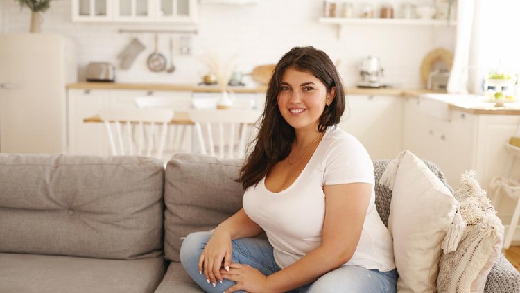 Relaxation, leisure, rest, domesticity and coziness. Beautiful charming young female with chubby cheeks and curvy body sitting on sofa barefooted, keeping one foot on floor, smiling at camera